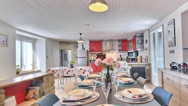 Un comedor con una mesa dispuesta para cuatro personas, un jarrón con flores y una cocina al fondo.