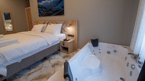 Une chambre d’hôtel avec un lit, une tête de lit en bois, un tableau représentant un paysage de montagne, une petite table de chevet avec une lampe et une grande baignoire.