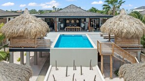Une piscine entourée d’une terrasse en bois, de constructions à toits de chaume et offrant une vue sur le paysage environnant.