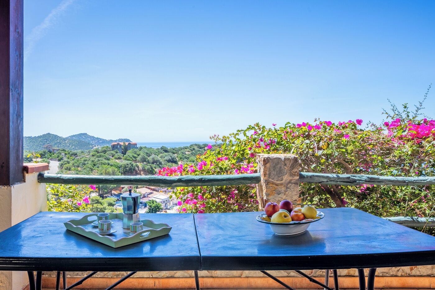 A balcony with a table, a bowl of fruit, and a view of a coastal landscape.