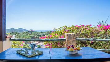 A balcony with a table, a bowl of fruit, and a view of a coastal landscape.