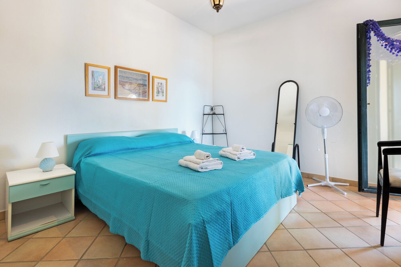 A bedroom with a bed, a nightstand, a wall-mounted shelf, a standing fan, and a door with a decorative item.