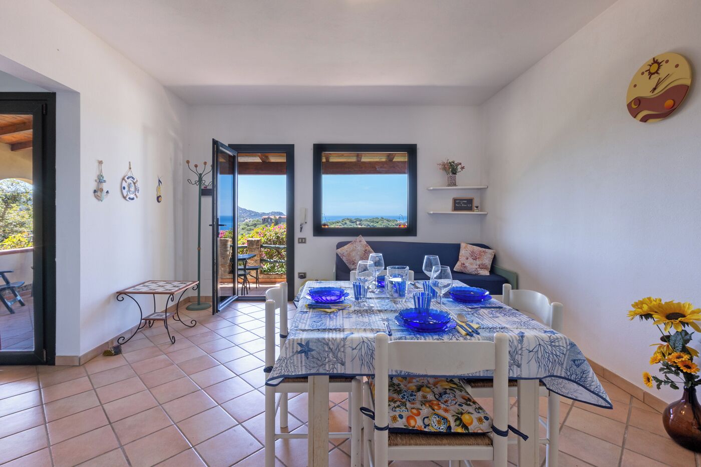 A dining area with a table set for four, a sofa, and a view through an open door to a balcony.