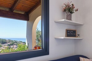 Una habitación con vista al mar, una ventana con una viga de madera, una repisa con un jarrón de flores y un cartel de bienvenida.