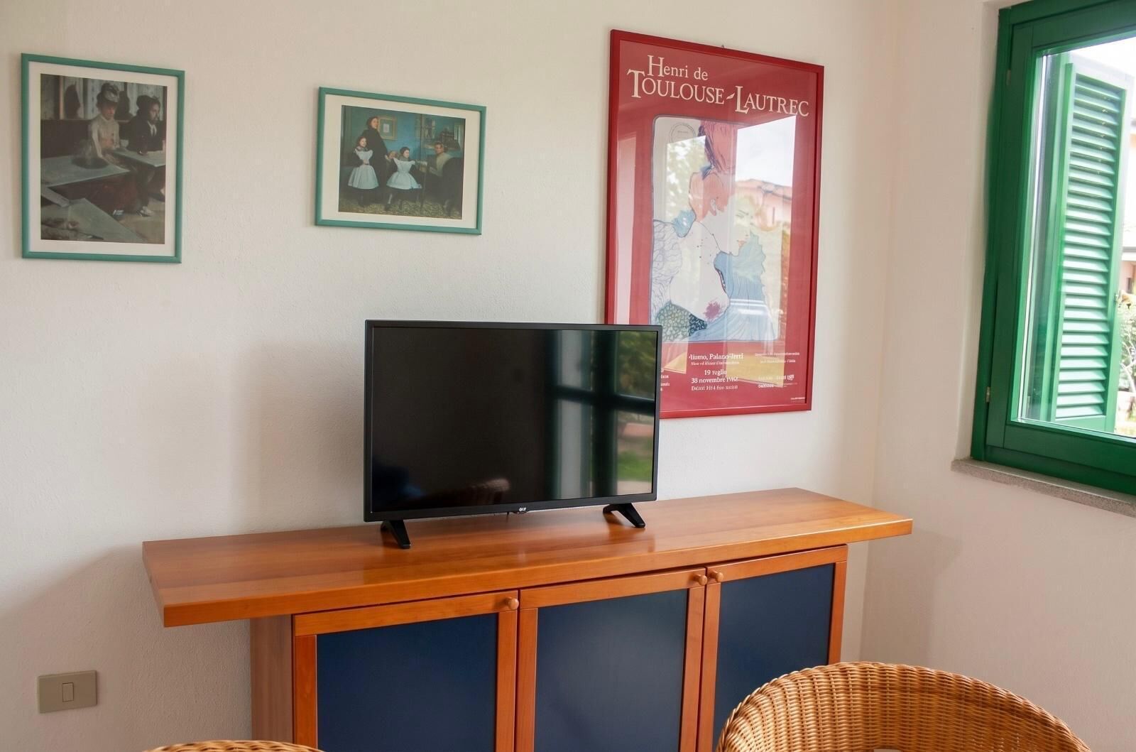 Een televisie op een houten standaard, twee ingelijste foto's aan de muur en een raam met groene luiken.