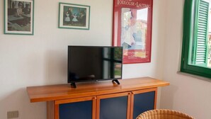 A television on a wooden stand, two framed pictures on the wall, and a window with green shutters. - Acqua del Mare | Welcomely (Tanaunella)