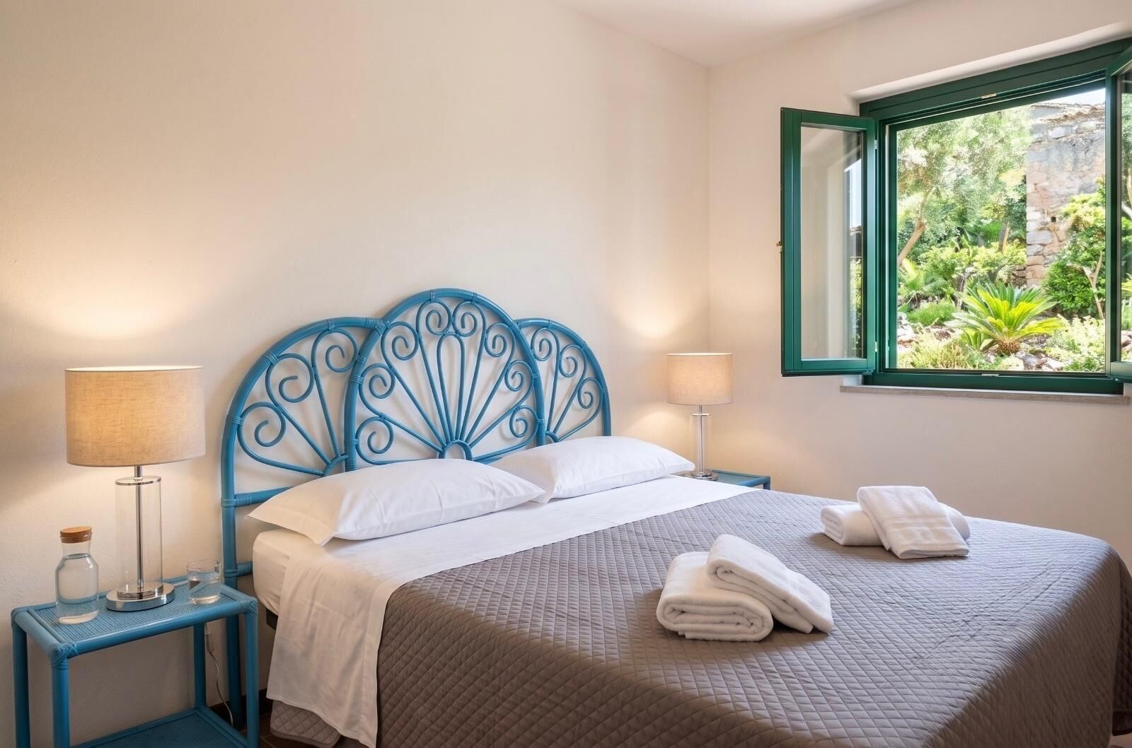 A bedroom with a bed, two bedside tables, lamps, a window with a view of greenery, and a decorative headboard.