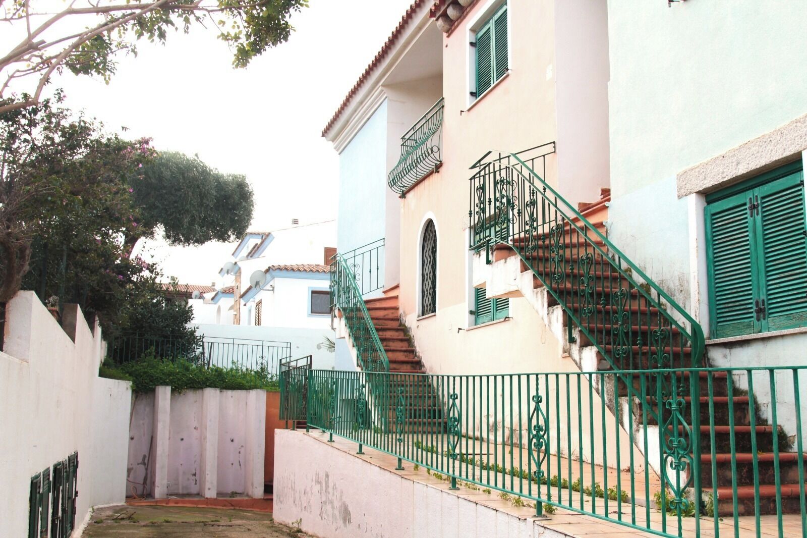 Uma rua estreita com um edifício branco, destacando-se uma escadaria com guarda-corpos verdes e uma varanda.