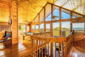 A wooden cabin interior with a pool table, a ceiling fan, and large windows offering a view of the outdoors. - Panoramic Views w/ Hot Tub, Pool Table, Arcade! (Sevierville)