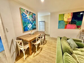 A dining area with a wooden table and white chairs, a green sofa, and two large abstract paintings on the walls. - Booyun@Bruns - 2 bedroom apartment - walk to shops, river and beach (Brunswick Heads)