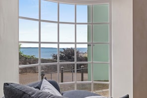 Ein Zimmer mit einem großen Erkerfenster mit Blick auf eine Küstenlandschaft, einer Steinmauer und einem gepflasterten Weg.