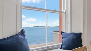 Ein Zimmer mit großem Fenster und Blick auf das Meer sowie eine entfernte Insel. Auf einer weißen gepolsterten Bank liegen zwei dunkelblaue Kissen.