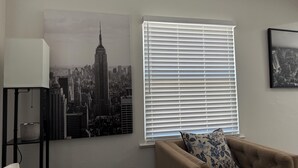 Ett rum med en stor målning som föreställer en stadsbild, ett fönster med persienner, en lampa och en soffa med en mönstrad kudde.