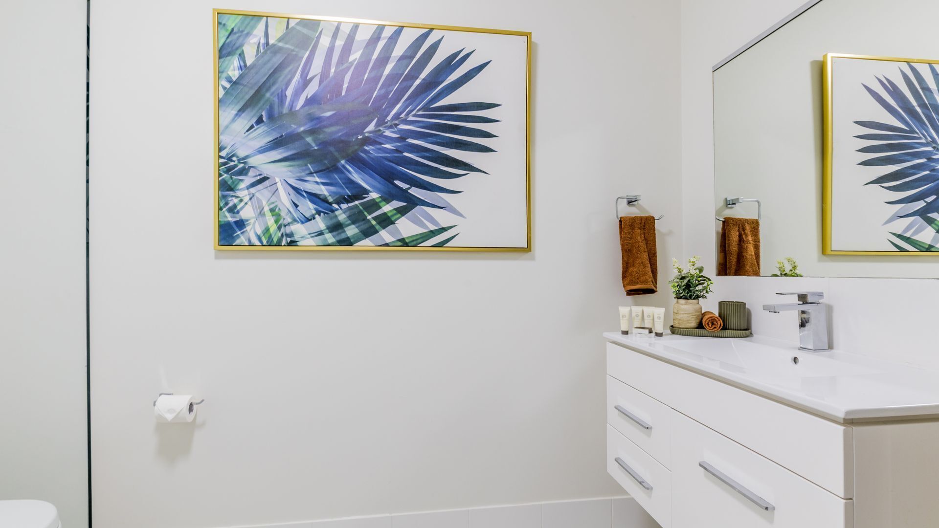 Un baño con tocador blanco, un espejo grande y una obra de arte con hoja tropical enmarcada.
