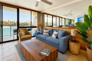 A spacious living room with a large sofa, a coffee table with a jigsaw puzzle, a wicker chair, and a view of the beach through large windows.
