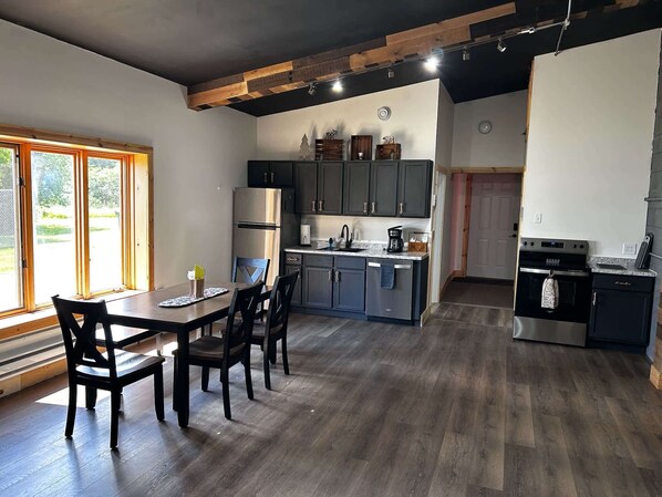 A modern kitchen with dark cabinets, a stainless steel refrigerator, and a dining area with a wooden table and chairs. - North Shore Den on 61 (Two Harbors)
