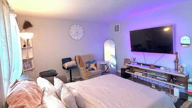 Ett hotellrum med en säng, ett skrivbord, en stol, en tv och en väckarklocka på väggen.