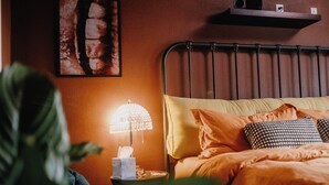 Makuuhuone, jossa on sänky, tyynyjä, yöpöytä lampun kanssa ja seinälle kiinnitetty hylly.