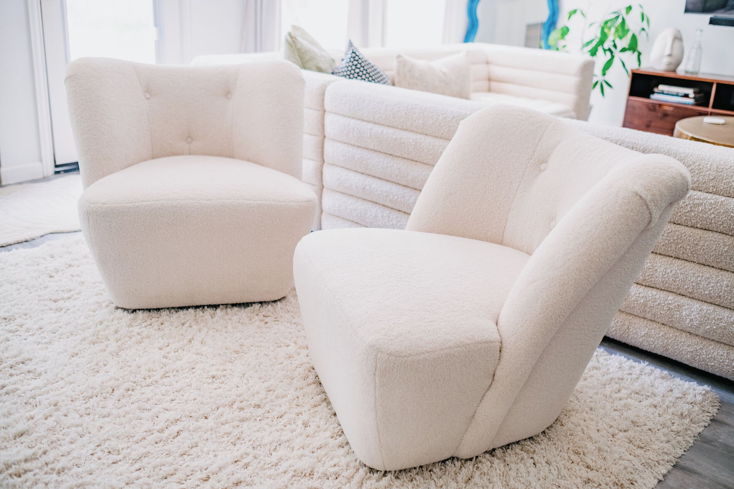 Uma sala de estar com sofá e poltronas brancos, um tapete felpudo branco e uma estante com livros e objetos decorativos.