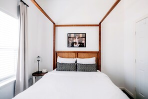 Een slaapkamer met een bed, nachtkastje, lamp, een lijst met foto aan de muur en een raam met gordijnen.