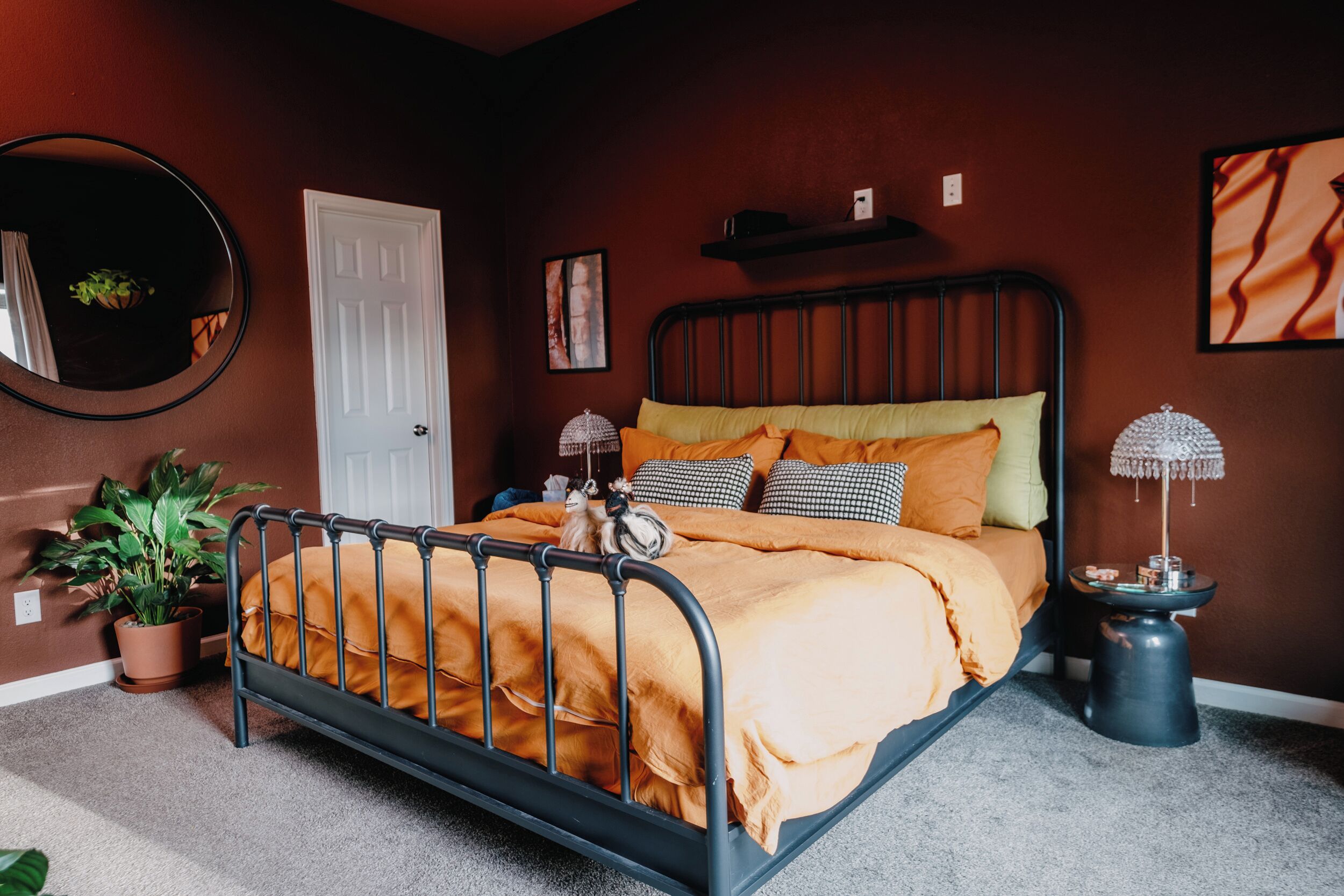 Quarto com estrutura de cama de metal, porta branca, espelho redondo, planta em vaso, mesa de cabeceira com abajur e prateleira na parede.