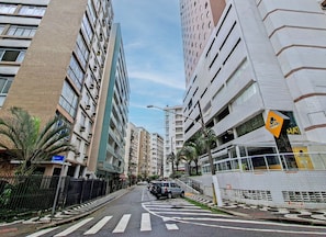 A street view with tall residential buildings, a pedestrian crossing, and a sign for 'The Hat' restaurant. - SHB - Flat vista mar no Boqueirão (Santos)
