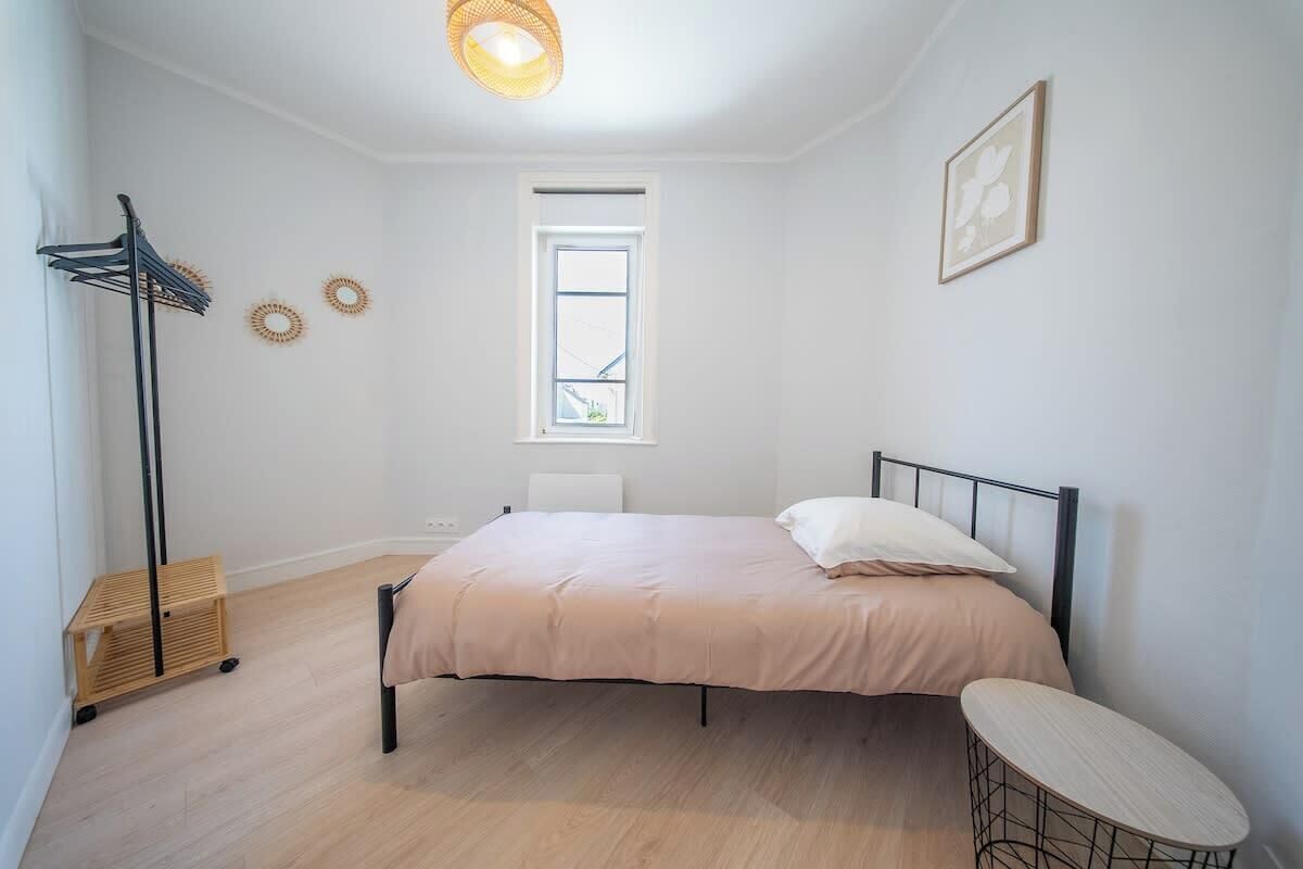 Ein Schlafzimmer mit einem Bett, einem Nachttisch, einer Stehlampe, einem Fenster und einem Wandbild.
