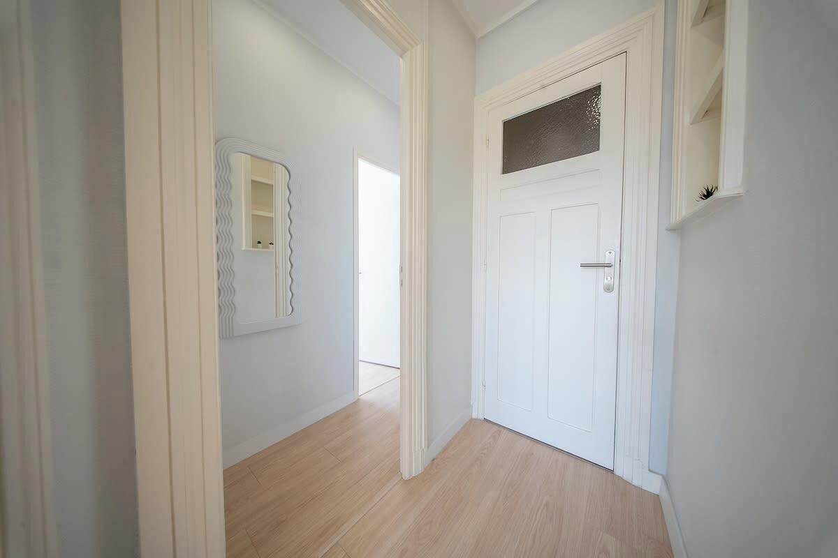 Een gang met een witte deur, een spiegel en een licht houten vloer.