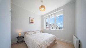 A bedroom with a large window, a bed with a patterned bedspread, a bedside lamp, and a small table.