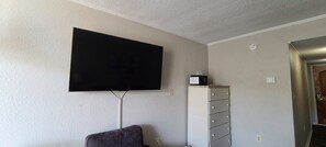 Una habitación con una televisión de pantalla plana montada, un sofá gris, un mueble con cajones y una puerta que da a otra estancia.