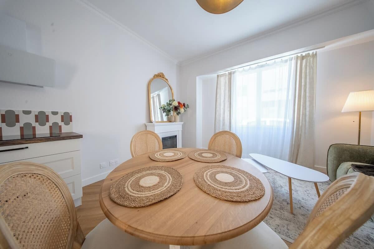 A dining area with a round wooden table, four woven placemats, wicker chairs, a white side table, a mirror, and a lamp.