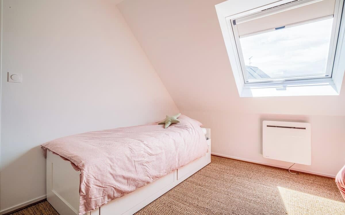 Une petite chambre avec un lit simple, un plafond en pente, une fenêtre et un climatiseur fixé au mur.