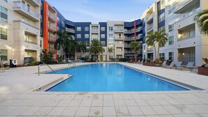 Una piscina con solárium empedrado, rodeada de edificios de varios pisos con balcones y palmeras.