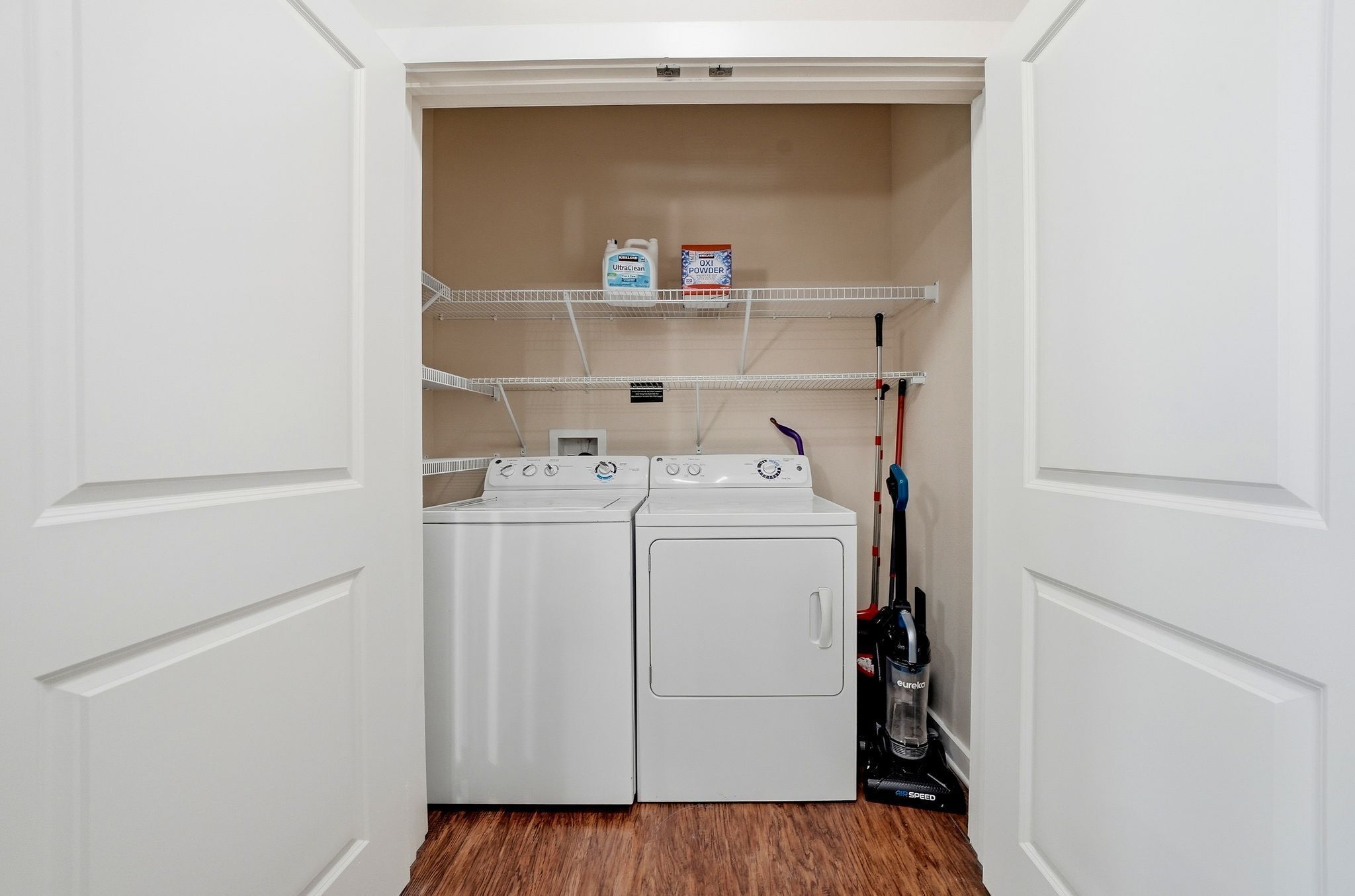 A laundry room with two white dryers, a shelf with detergent, and a vacuum cleaner.