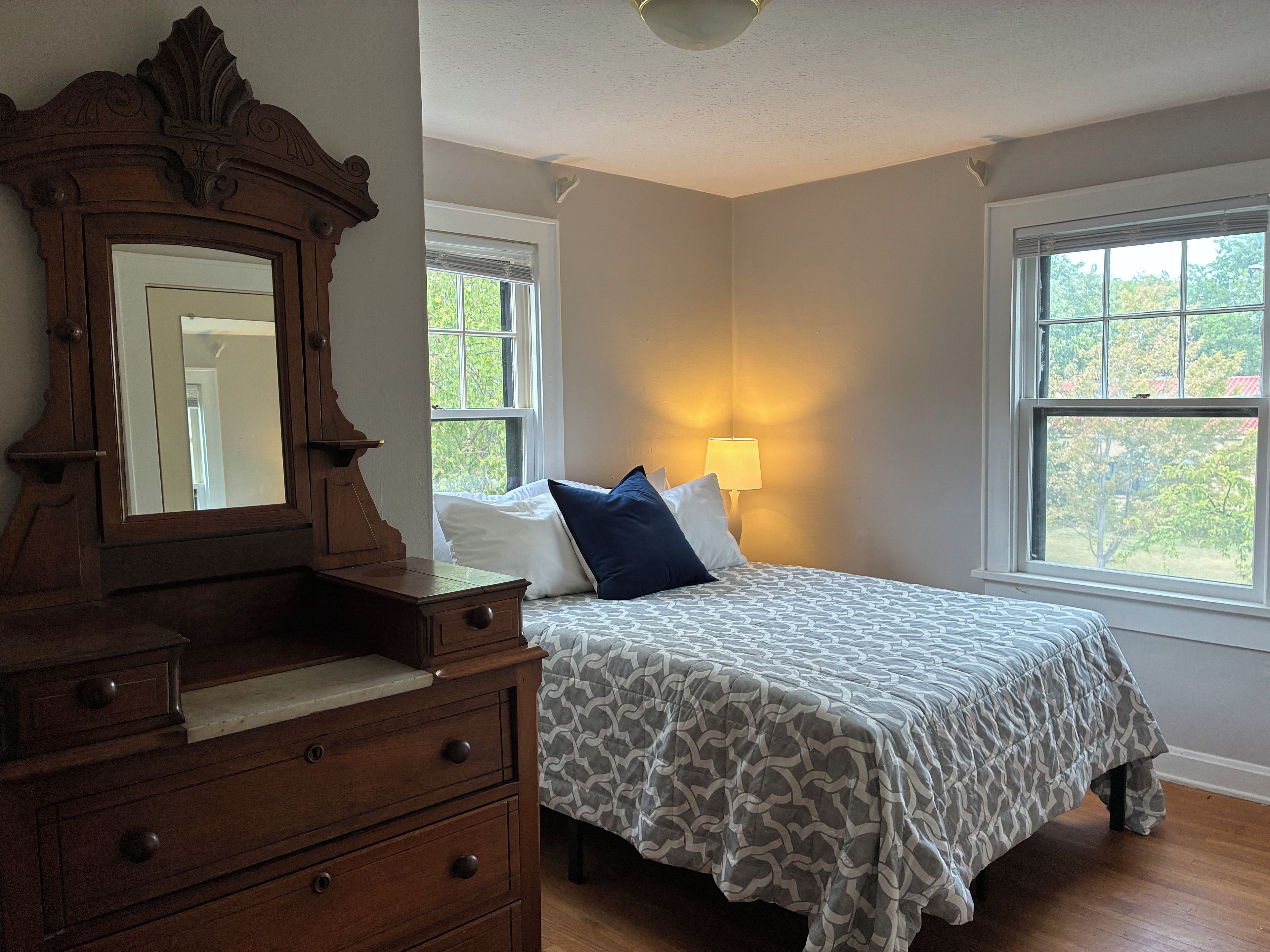 Un dormitorio con cama, cómoda, espejo, ventanas y lámpara.