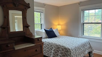 Un dormitorio con cama, cómoda, espejo, ventanas y lámpara.