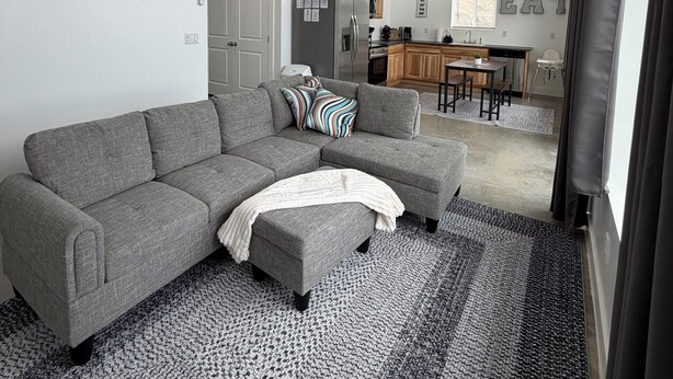 A modern living room with a grey sectional sofa, a ceiling fan, and a kitchen in the background.
