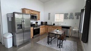 A modern kitchen with stainless steel appliances, wooden cabinets, a dining area with a table and chairs, and a 'EAT' sign on the wall.