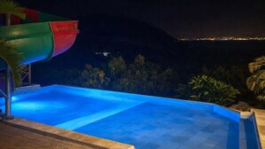 A nighttime swimming pool with a slide, surrounded by a stone deck and palm trees, overlooking a cityscape.