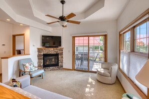 A living room with a stone fireplace, a ceiling fan, a television, and a sliding glass door to an outdoor patio. - Hilltop Escape - Enjoy Lake Superior Views with City Amenities (Bayfield)
