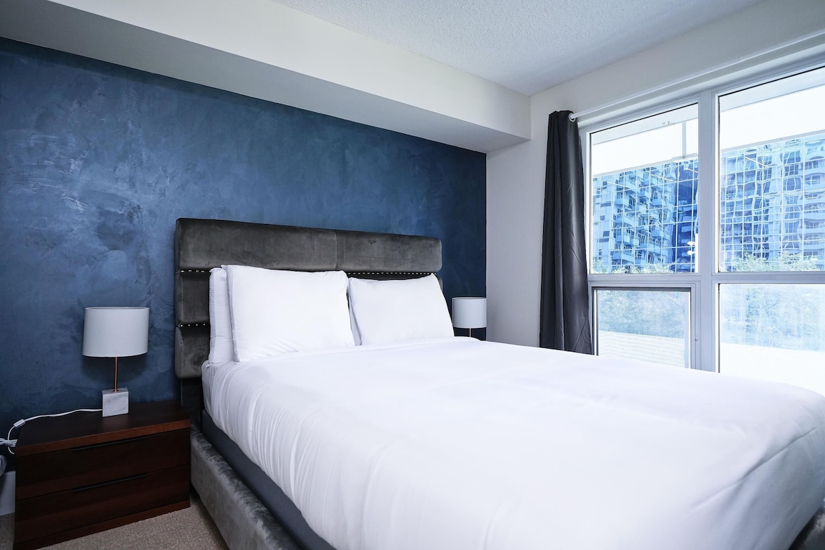A bedroom with a large bed, a nightstand, a lamp, and a window with a view of buildings.