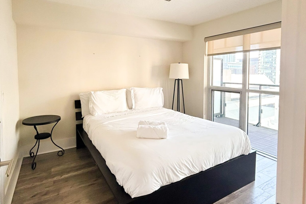 A hotel room with a bed, a small table, a floor lamp, and a large window with a view of buildings.