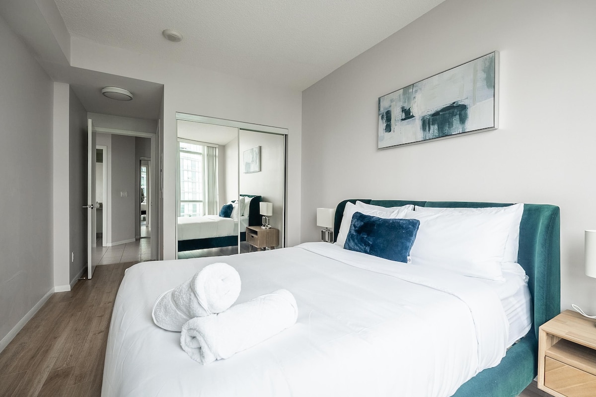 A modern hotel room with a large bed, a bedside table, a mirror, and a painting on the wall.