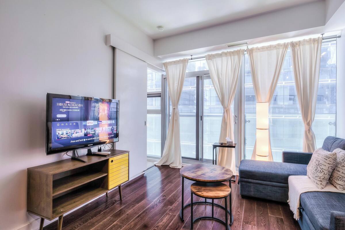 Una sala de estar moderna con una televisión de pantalla plana, muebles de madera, un sofá y ventanas grandes con cortinas.