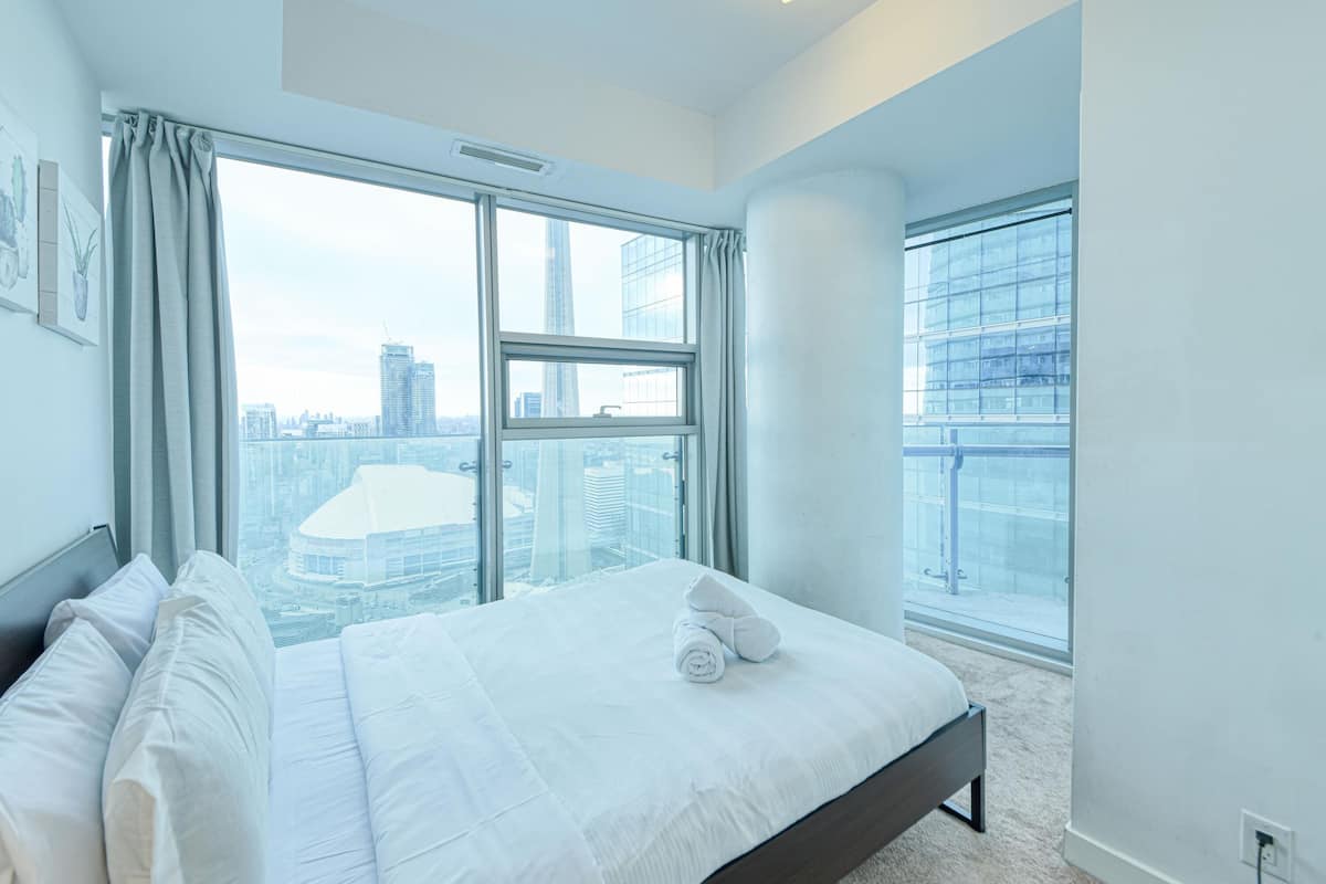 Una habitación de hotel moderna con una cama grande, vistas a la ciudad a través de ventanales del suelo al techo y un balcón con barandilla de cristal.