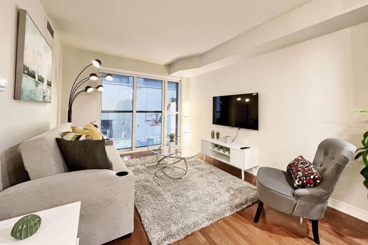 Una sala de estar moderna con un sofá y un sillón grises, una mesa de centro, un televisor montado en la pared y un ventanal con vistas al exterior.