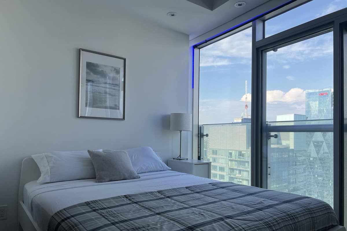Una habitación de hotel moderna con una cama grande, un cuadro en la pared y vistas a la ciudad a través de un ventanal amplio.