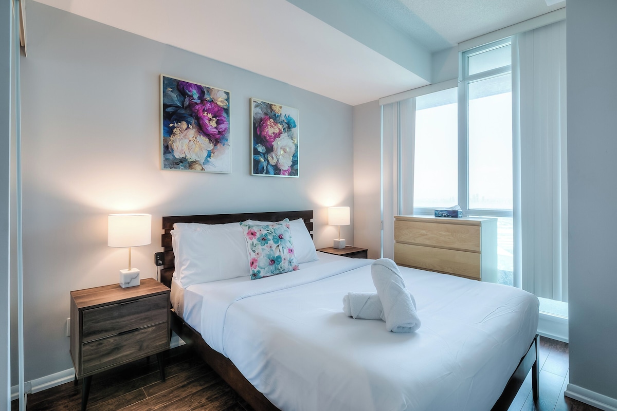 A hotel room with a neatly made bed, two wall-mounted paintings, a nightstand with a lamp, and a window with curtains.