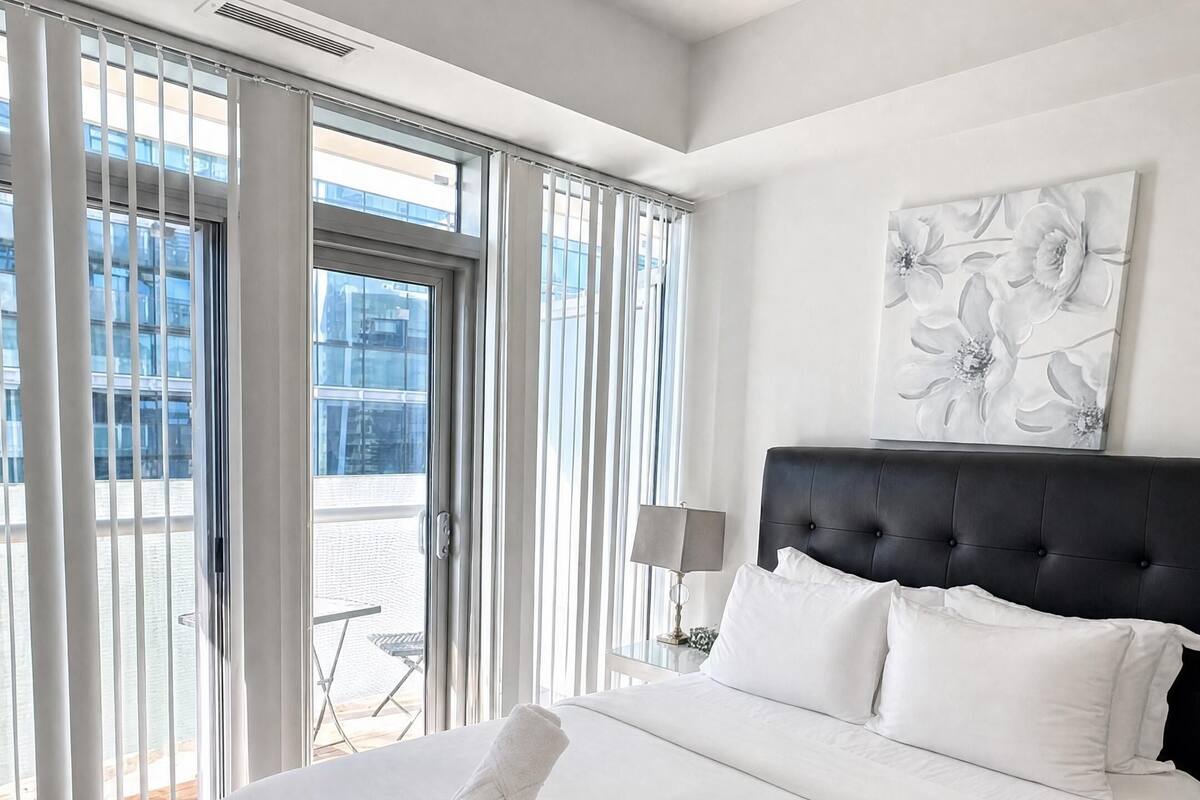 Habitación de hotel con una cama grande, cabecero, ventana con persianas, un cuadro en la pared y vistas al exterior.