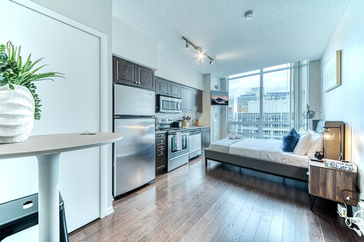 A modern apartment with a kitchen, a dining area, a bed, and a large window with a city view.